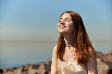 Fototapeta na wymiar A beautiful red-haired girl framed her face with the rays of the sun on the beach and smiles a wide white smile. 