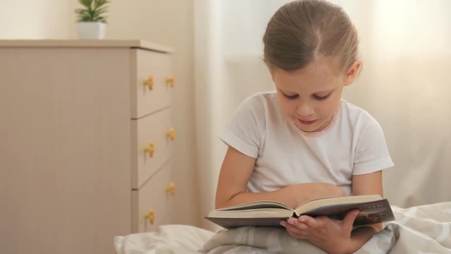 Adorable little girl holding story book, sitting on the bed and reading. 