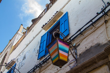 Fototapeta na wymiar Open window with blue shutters shutters against a white wall and blue sky with a bright multi-colored lantern