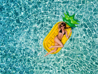 Sportliche, blonde Frau im Bikni treibt auf einer aufblasbaren Ananas über türkis blaues Wasser