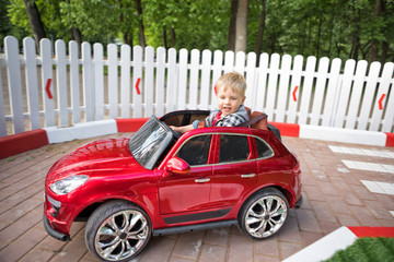 Little smiling boy is driving a car on kid's go-karting. Three year-old child in a red toy car in the children's race track with road signs and traffic light having fun outdoors, attention concept