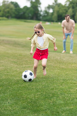 Obraz na płótnie Canvas adorable child in sunglasses playing with soccer ball while father standing behind in oark