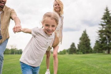 Fototapeta premium cropped shot of parents with daughter holding hands and child smiling at camera in park