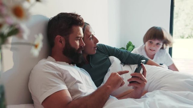 Lovely married couple lies in bed in pajamas using their phone, their cute little son and daughter join them, dad is taking selfie of the whole family. Perfect morning, happiness, modern parents.