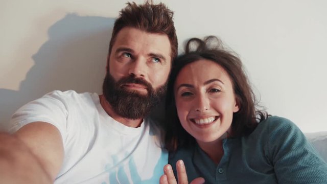 Smiling bearded man with his pretty young wife recording video for vlog while staying in bed, recommending, big thumb up, smiling. Modern lifestyle, being online, internet. Contemporary families.