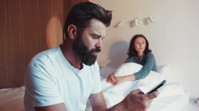 Sad bearded man in white shirt sits on the corner of the bed, using the smartphone, his unhappy wife sits on the background. Jealousy, conflict situation, no trust. Fighting, arguing.