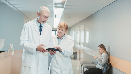 In the Hospital Two Doctors Walking Through the Hospital Hallway, They Talk and Share Tablet Computer. Modern Hospital with Patients and Medical Personnel.