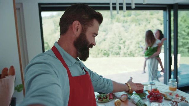 Attractive bearded vloger records room-tour video about his modern light kitchen, tastes sweet red pepper, invites his wife with a cute child to greet the subscribers. Food vlog, modern parents.