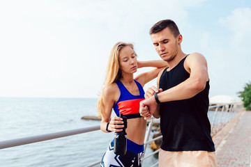 Muscular handsome man and sportive girl checking a time on smart watches, after running workout on the quay, near the sea. Sport concept.