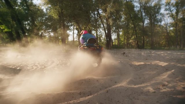Man riding atv on sand picking up pillar of dust at sunset. Modern guy driving quadbike at extreme holiday. Tourist man driving quad bike on sand in slow motion. Extreme driving sports at forest