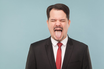 Closeup portrait of crazy man in classic suit, tongue out, and roar. indoor studio shot. isolated on light blue background. handsome businessman with black suit, red tie and mustache with closed eyes