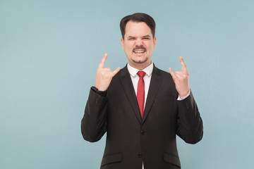 I'ts rock and roll, yeah! Expressive cool businessman showing rock sign. indoor studio shot. isolated on blue background. businessman with black suit, red tie and mustache looking at camera.