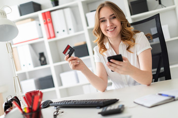 Beautiful young girl sits in the office, holds a bank card and phone in her hand.