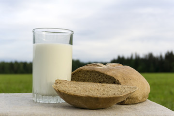 a glass of milk and a loaf of rustic bread on the corner of the table with a plain linen tablecloth against the background of a blurred meadow