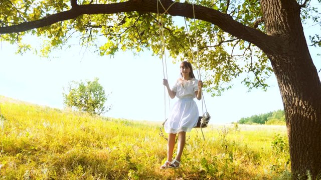 Beautiful girl with long hair swings on swing under summer oak in white dress.