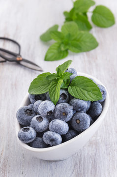 Drops fresh blueberries in a white bowl, with meant leaves on a wooden background