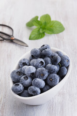 Drops fresh blueberries in a white bowl, with meant leaves on a wooden background