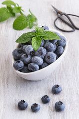 Drops fresh blueberries in a white bowl, with meant leaves on a wooden background