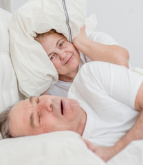 Irritated senior woman blocking ears with pillow while her husband snoring on bed