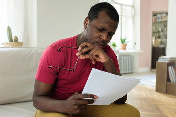 Photo of young African American man sitting on couch at home holding documents or bills, having taken off glasses, looking deeply troubled because of high expenses and facing problems with budget
