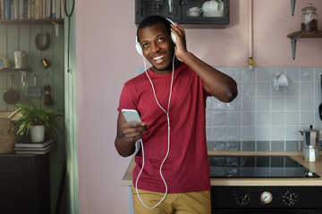 Horizontal shot of African American man standing in his kitchen in casual clothes with big headphones on, listening to music from his phone smiling happily and enjoying his free time at home