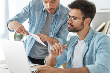 Obraz premium Prosperous men colleagues dicusss business project while look attentively in documents, work with modern laptop computer, talk about working strategy of cooperation. Office work and collaboration