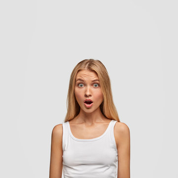 Vertical shot of stunned blonde young female in casual white vest, hears horror news or something bad, isolated over white studio background, keeps jaw dropped. People and astonishment concept