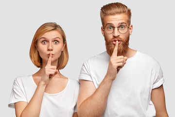 Horizontal shot of two girlfriend and boyfriend make silence gesture, ask not tell about their...