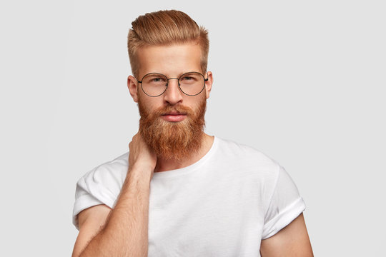 Horizontal shot of handsome serious male boss has trendy haircut and ginger beard, keeps hands behind neck, looks confidently at camera, prepares for meeting with colleagues, isolated on white