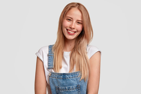 Glad European young female with satisfied expression, smiles broadly, has healthy pure skin, dressed in denim stylish overalls, being in god mood after walk in park, isolated over white background