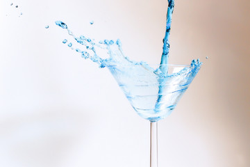 Cocktail with blue liquid in glass. Glass with blue water pouring with liquid with splashes and drops. Martini glass filling with alcohol with splashes on white background. Refreshing drink concept