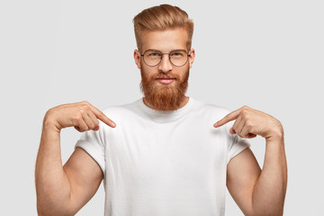 Horizontal shot of confident ginger male with trendy haircut, dressed in casual t shirt, indicates with both fore fingers at blank space for your logo, isolated over white background. Clothing concept