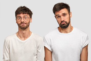 Fototapeta na wymiar Puzzled sad two bearded young guys stand closely to each other, express negative emotions, look in bewilderment, wear white casual clothes, have some troubles with business. People and emotions