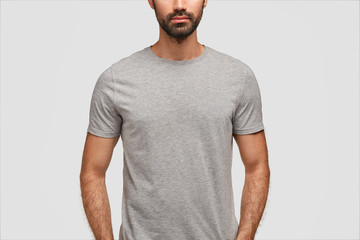 Young bearded male wears casual gray t shirt with blank space for your design, advertisement or promotional text, enjoys high quality textile, isolated over white background. Clothing concept