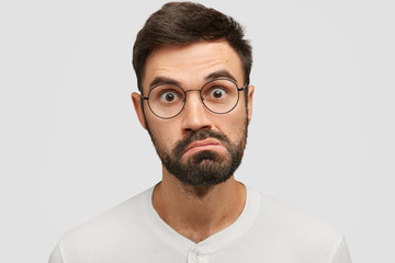 Obraz premium Close up portrait of handsome angry surprised Caucasian male looks in bewilderment, purses lips and stares at camera, wears glasses, isolated over white background. People, facial expressions