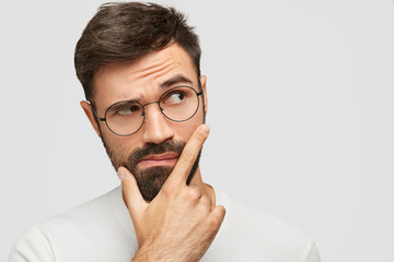 Headshot of good looking bearded young male holds chin and looks pensively aside, raises eyebrows...