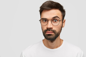 Emotive bearded young guy with thick stubble, trendy hairdo, looks with surprisement at camera, stares through glasses, dressed in casual white t shirt, expresses bewilderment and astonishment