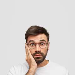 Vertical shot of stupefied Caucasian male touches cheek and stares at camera, realizes somebody told him lie, wears round spectacles, isolated over white background. People and reaction concept