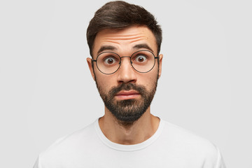 Omg concept. Surprised astonished young man stares with bugged eyes, can`t believe in his failure, has dark thick stubble, stands against white background. People, surprisement, shock concept
