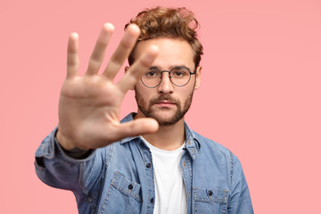 Serious handsome bearded hipster stretches hand towards camera, shows palm, demonstrates stop or...