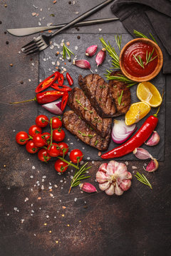 Grilled beef steaks with spices and vegetables on slate board, dark background