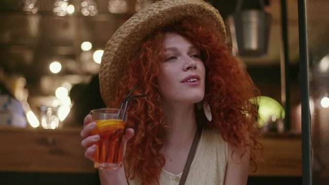 Stylish young attractive pretty woman with red hair in hat sitting in cafe waiting drinks a cocktail fashion girl connection summer portrait caucasian smartphone lady close up slow motion