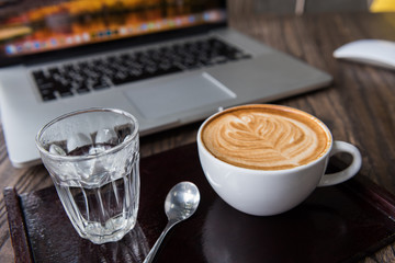 A Cup of hot heart latte art coffee on wooden plate with laptop computer