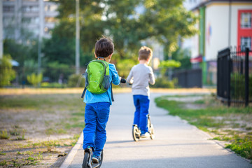 Two cute boys, compete in riding scooters, outdoor in the park, summertime. Kids are happy playing outdoors. Two brothers standing on their scooters