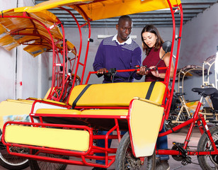 Polite African-American bikecab driver talking to young woman, offering rickshaw service