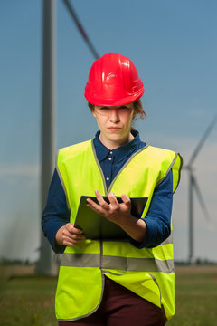Portrait of a serious successful young female engineer in a green vest and red hard hat designing a plan against a background of windmills