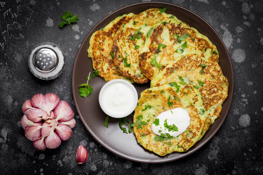 Zucchini pancakes with parsley, garlic and sour cream, summer food, tasty snack. In a dark plate on a dark background