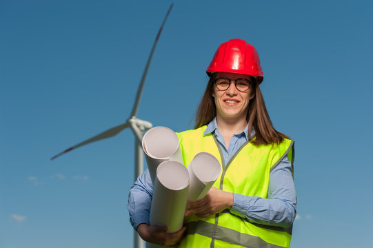 Portrait of a positive cute young female engineer in a green waistcoat and red hard hat with geodesic plans in hand against the backdrop of a windmill and blue sky.