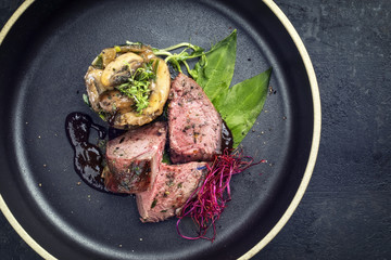 Traditional barbecue dry aged sliced venison fillet steak with potato mushroom terrine and wild garlic as top view on a plate with copy space