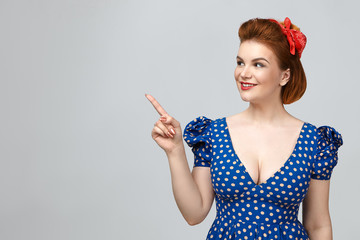 Obraz premium Beauty, style, fashion and elegance concept. Pretty girl dressed in pin up vintage style showing copy space for your information, pointing fore finger at blank studio wall and smiling broadly